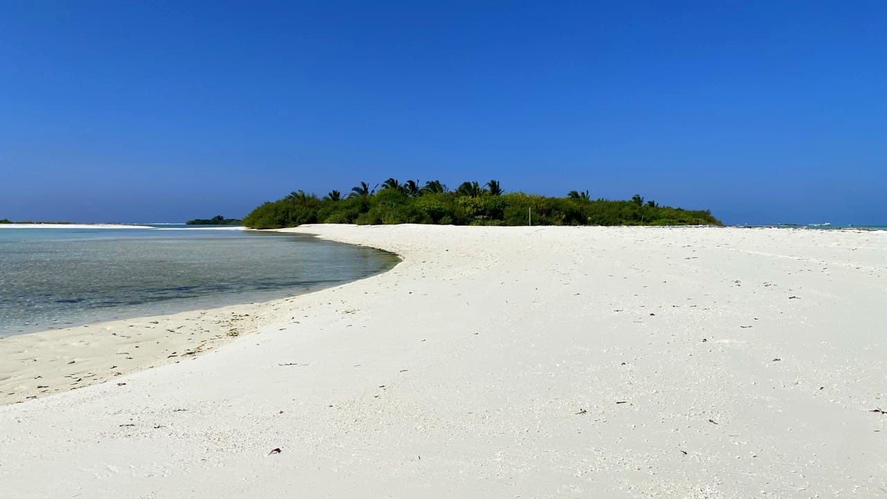A white sandbank curving across a turquoise lagoon toward a small palm-covered island under a deep blue sky